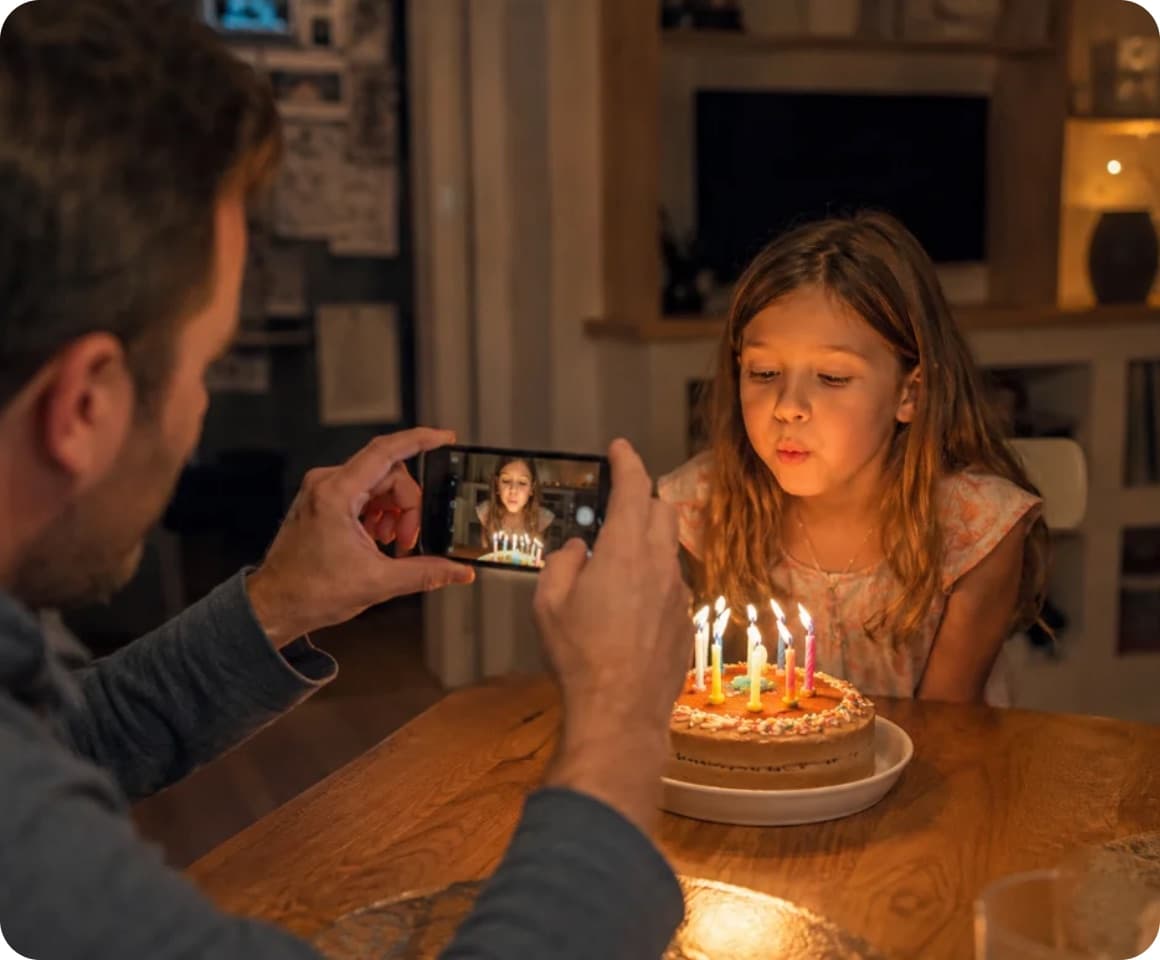 A father filming his daughter blowing out birthday candles in warm home light.